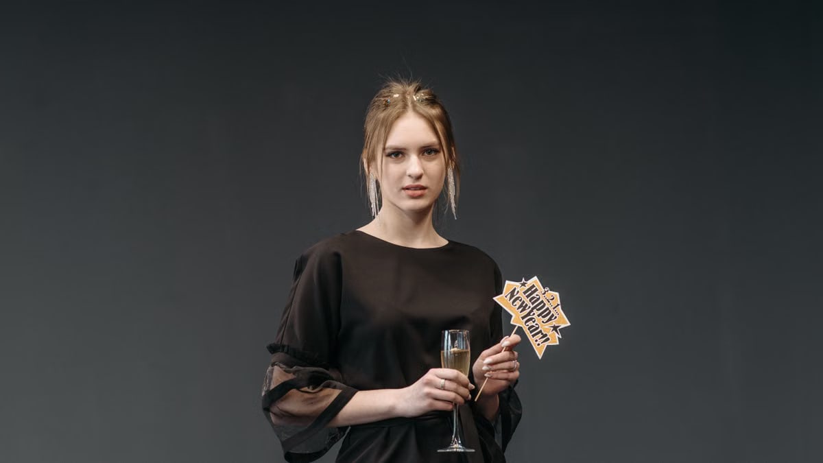 Caucasian woman in black dress holding champagne and a New Year sign, studio setting.